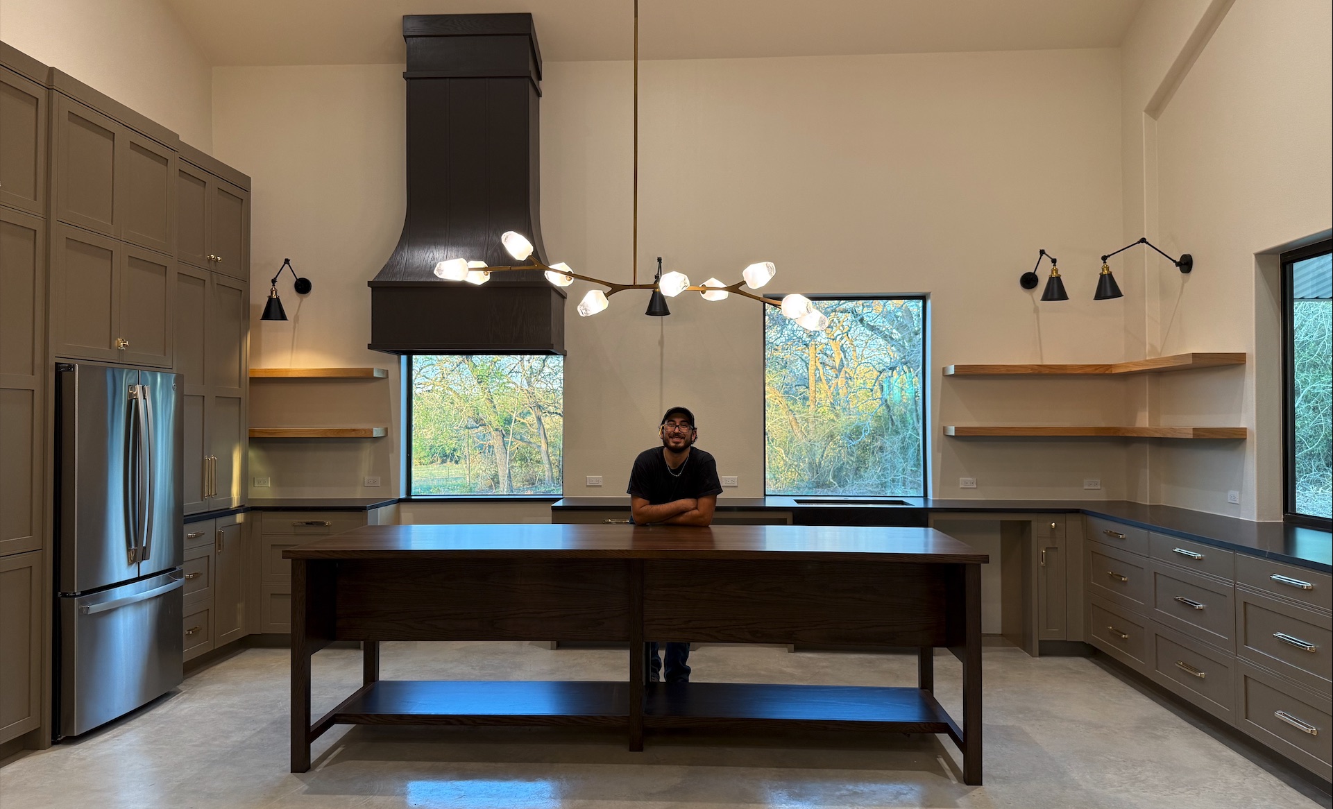 The maker behind Triple L Woodworks leaning on a large wood island in a finished kitchen with custom cabinetry and open shelving