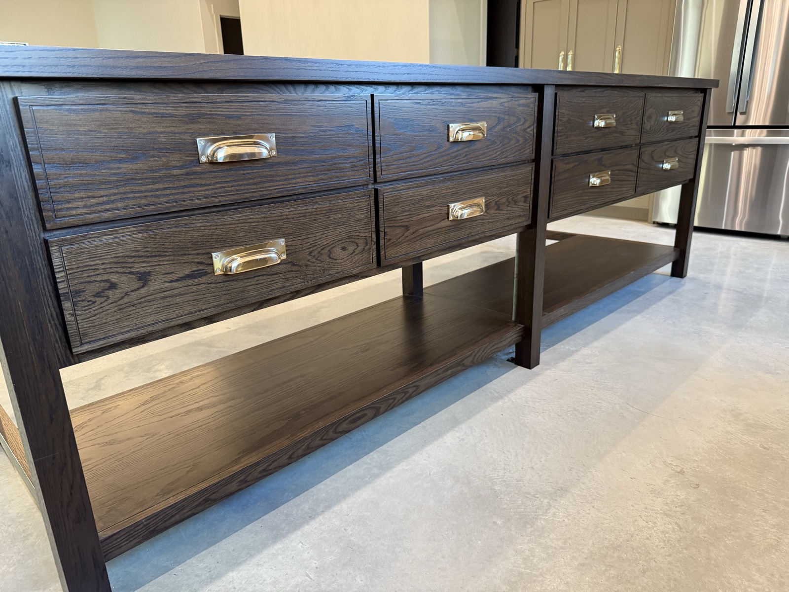 Angled view along the dark oak island showing its drawers, brass cup pulls, and lower shelf