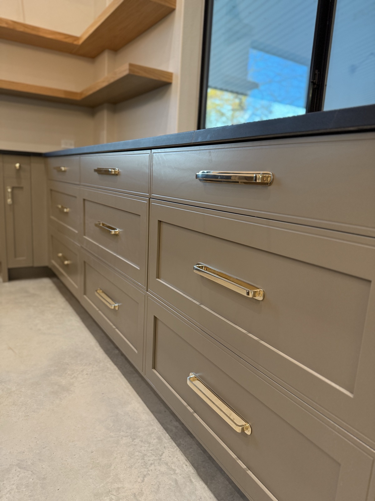 Run of greige shaker drawers with long polished brass pulls under a dark countertop and natural wood floating shelves