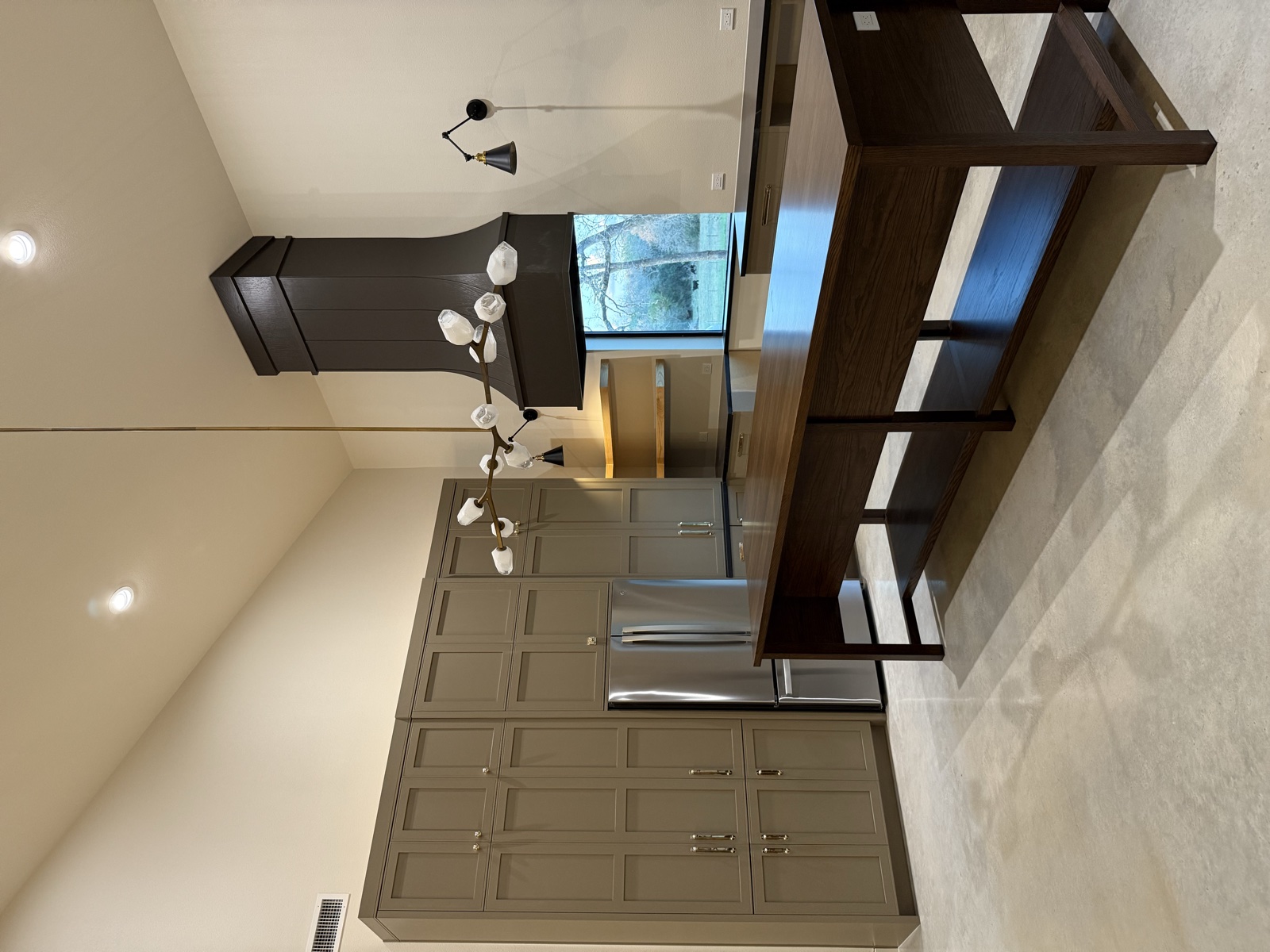 Kitchen with a dark-stained oak island on open legs, a tall wall of greige shaker cabinets around the refrigerator, and a dark wood range hood under a vaulted ceiling