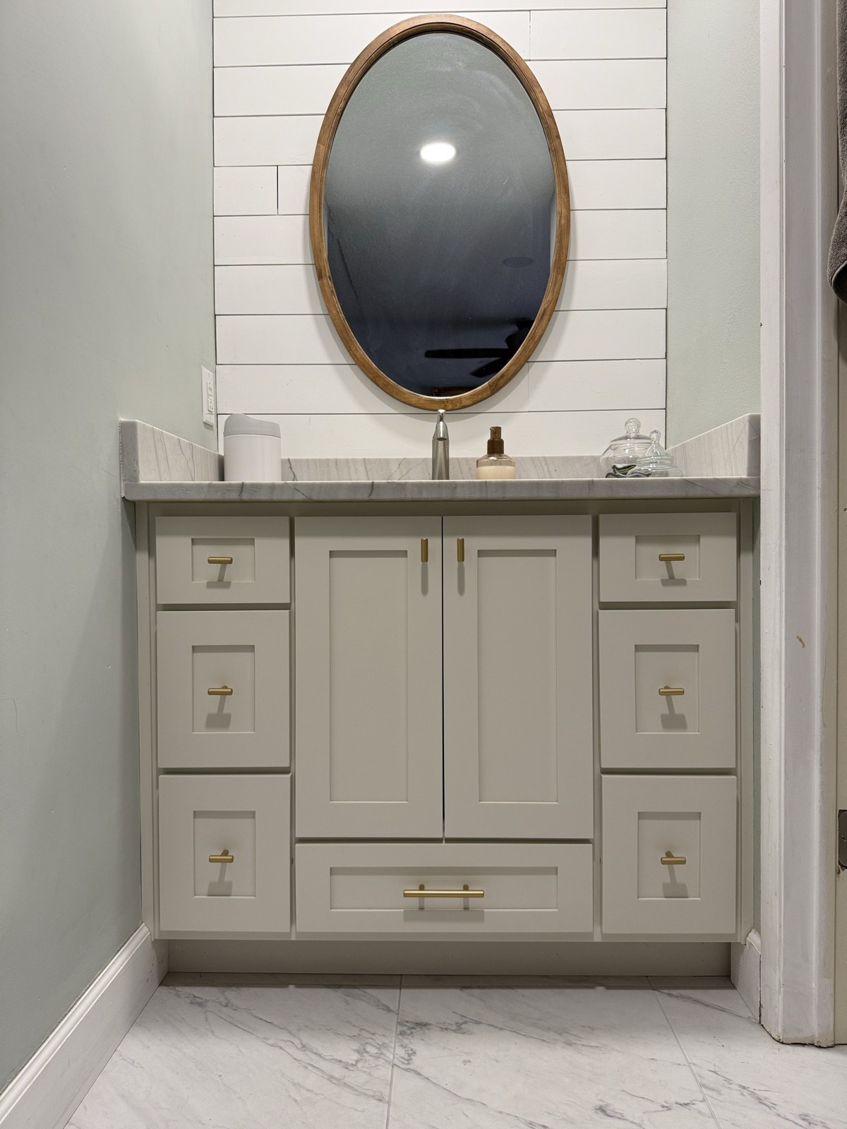 White shaker bathroom vanity with banks of drawers and brass pulls, under an oval wood mirror on a shiplap wall