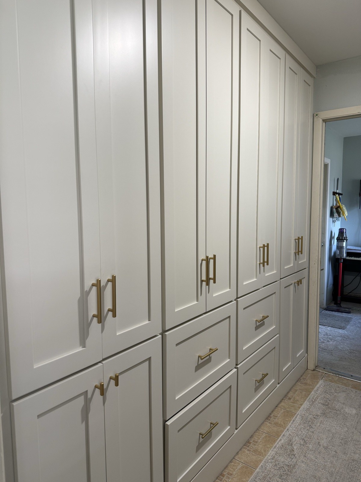 Wall of tall white shaker pantry cabinets and drawers with brass pulls lining a hallway