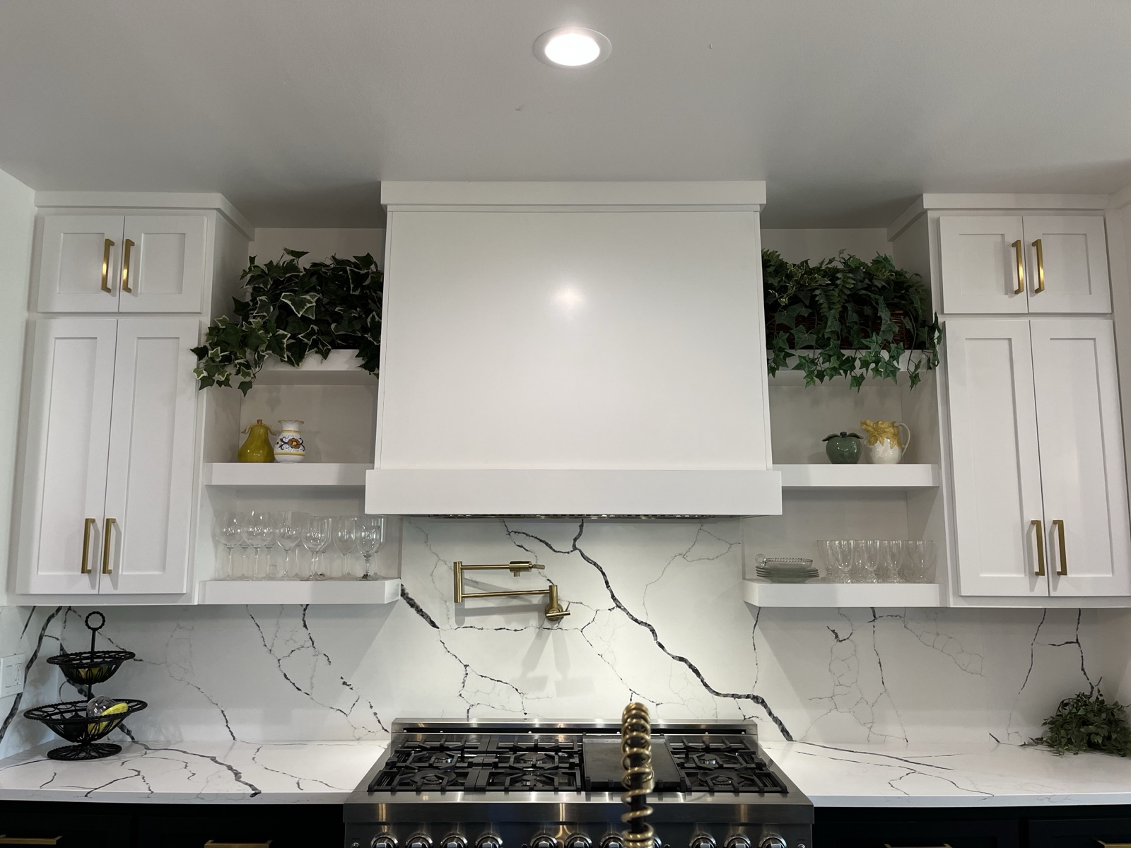 White range hood flanked by open floating shelves and shaker cabinets with brass pulls above a marble-look backsplash